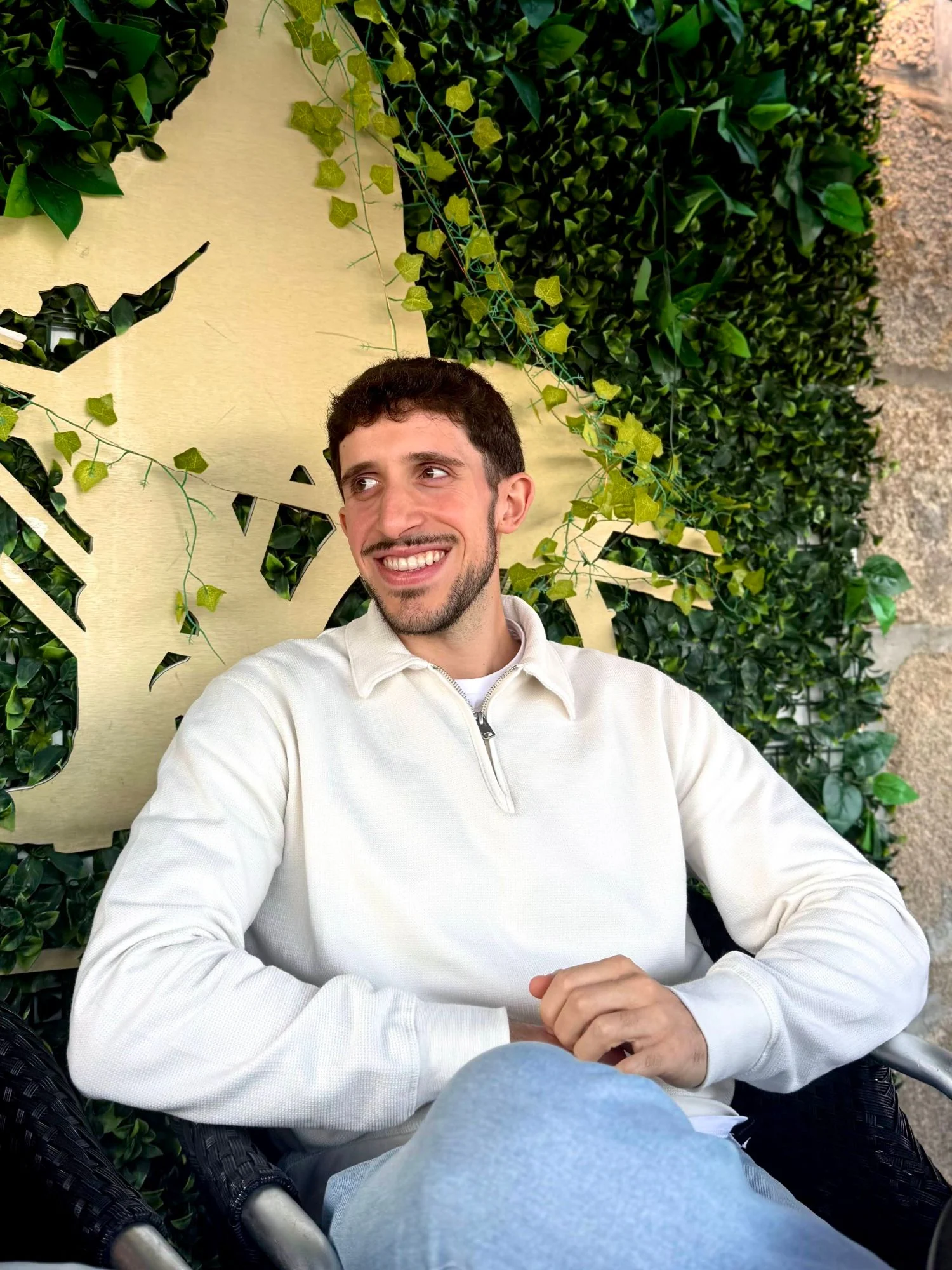 Gonçalo Silva, smiling, in a cream half-zip sweater, seated in front of a green plant wall